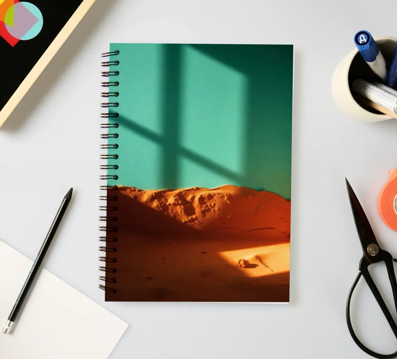 sunlight casts geometric shadows on orange sand and turquois spiral notebook by DesignDoodle