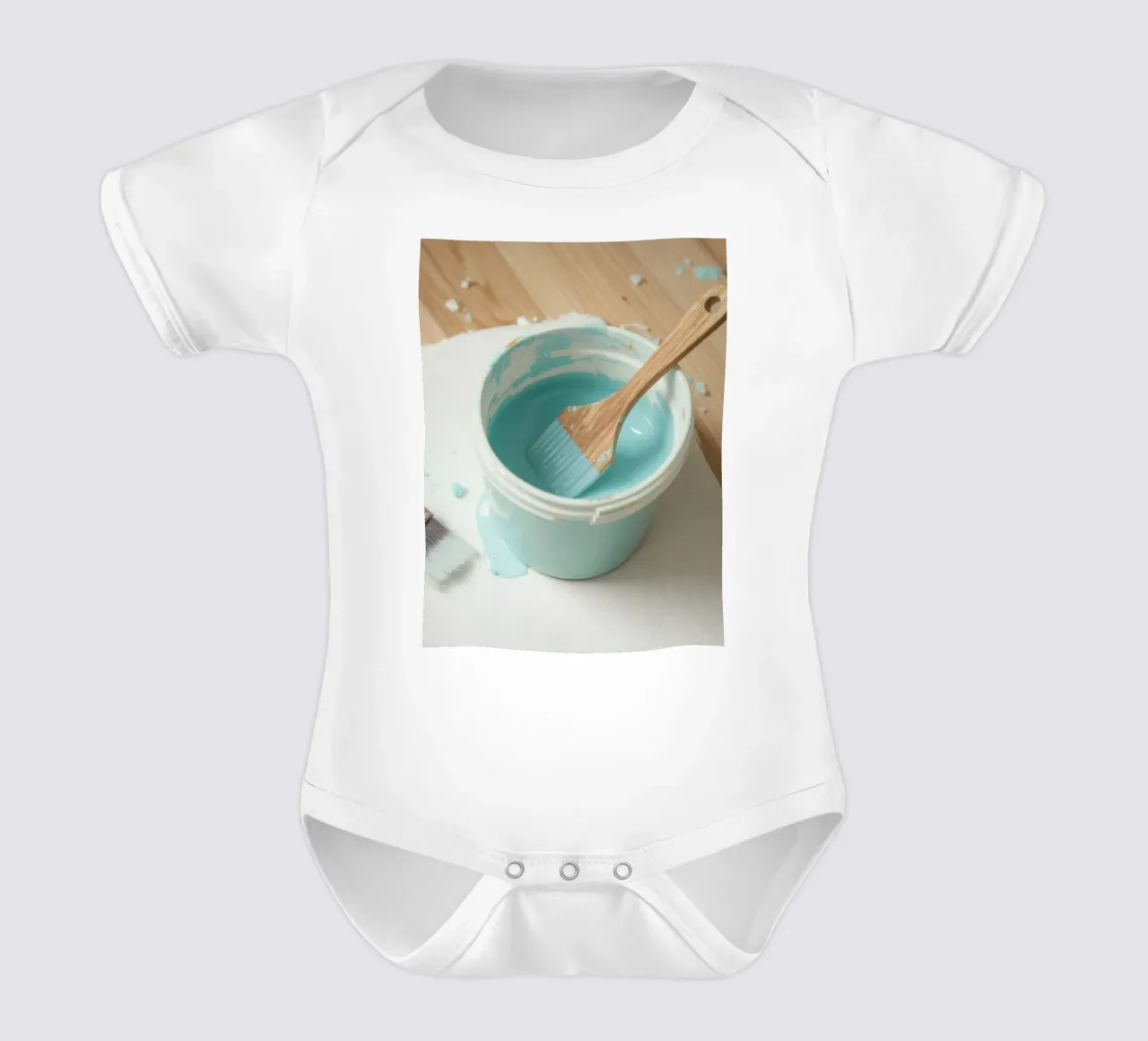 Un pincel de madera descansa en un bote de pintura azul junto a una brocha pequeña body de bebé de DesignDoodle